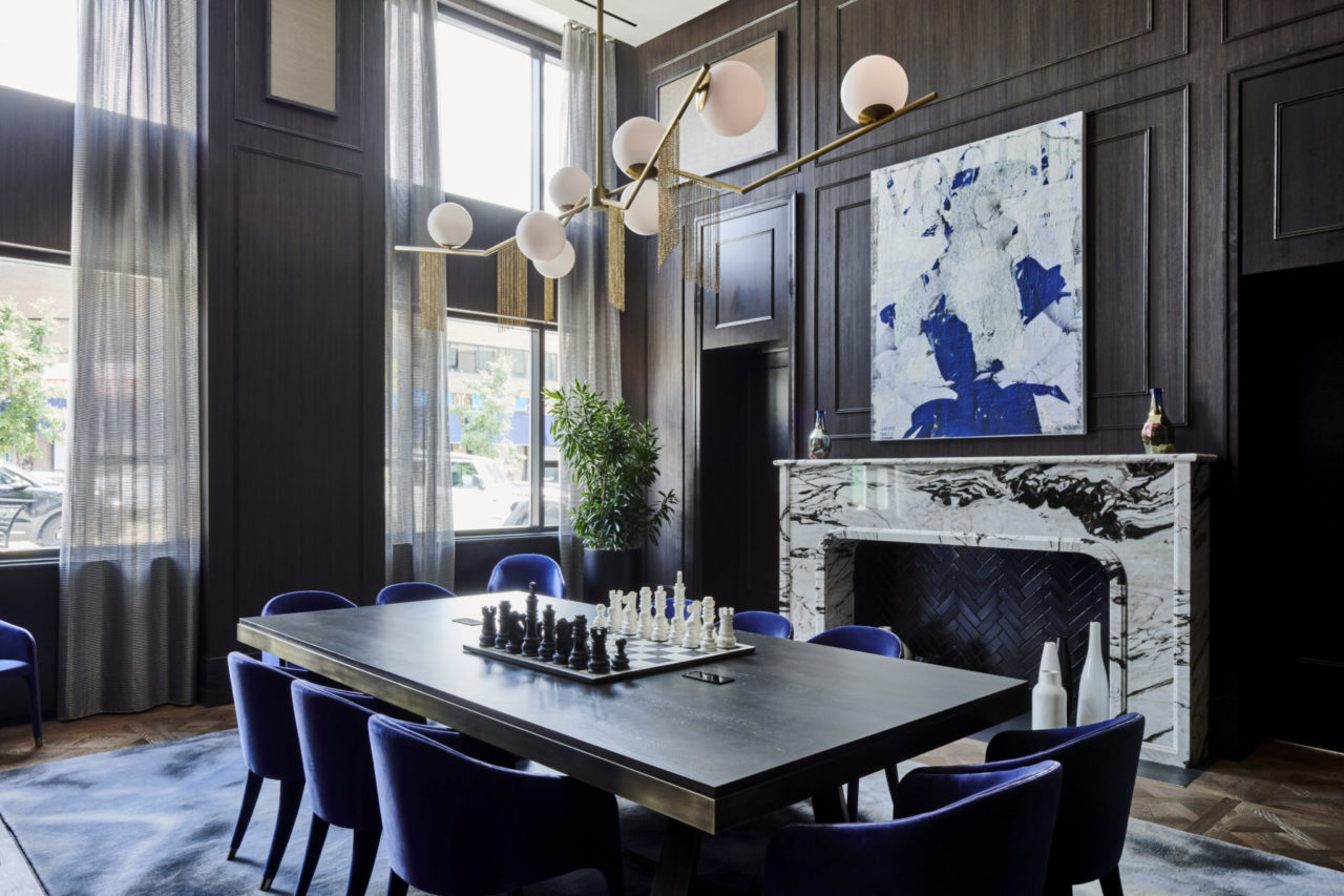 Elegant blue chairs surrounding a large table with a chess set on it with wood paneling & fire place at our hotel in Birmingham, MI